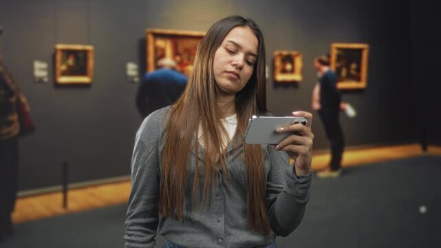 Young hispanic woman brunette with long hair holding smartphone, hand to face in building gallery with framed paintings and visitors; boredom distraction.