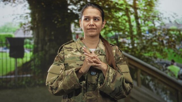 Woman soldier in camouflage uniform with hands interlaced at chest, standing by a wooden railing and tree on a street walkway; duty determination.