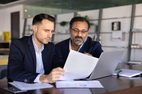 Two focused business men colleagues reviewing financial statements and revenue report, analyzing printed charts, referencing data on laptop. Project data analysis, analytics and team work in office