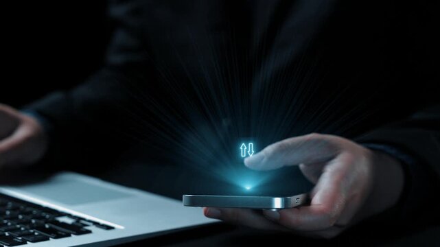 A businessman uses a smartphone to review quality and cost metrics while working on a laptop in a dark office. The image illustrates data analysis in a modern environment. Qubit
