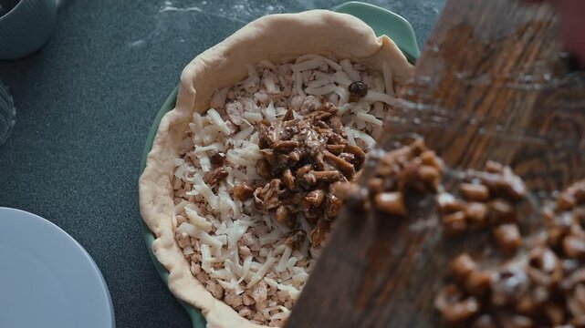 A person prepares a pie crust by adding a layer of shredded ingredients in a bowl. They then chop and add apple filling, taking care to ensure an even distribution for baking