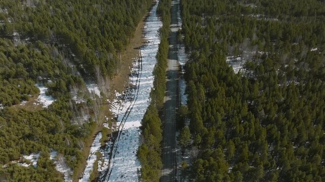 Drone footage of a straight asphalt road cutting through taiga forest with patches of melting snow in early spring, Buryatia, Siberia, Russia, top down view
