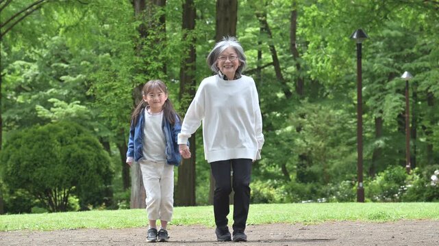 春の新緑の公園で遊ぶおばあちゃんと手を繋ぐ歩く寄り添う笑顔の孫娘のカメラ目線全身スローモーションの動画
