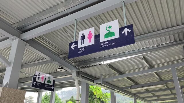 Bilingual signage in Malay and English directs users to restrooms and a prayer room. Simple icons ensure clarity. The sign hangs from a high structure for maximum visibility.