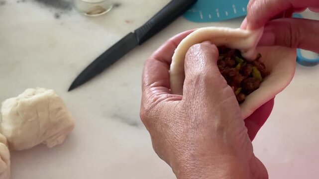 A close-up shows the hands of an elderly person pleating a handmade baozi, leaving a small opening at the top that reveals the savory minced meat and petai stuffing inside.