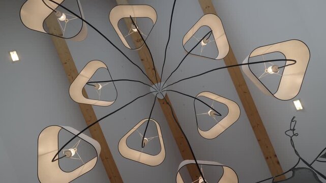 slow push out rotating shot of a modern multi-arm chandelier with rounded triangular fabric shades and exposed black cables radiating from central mount on white ceiling with wooden beams.