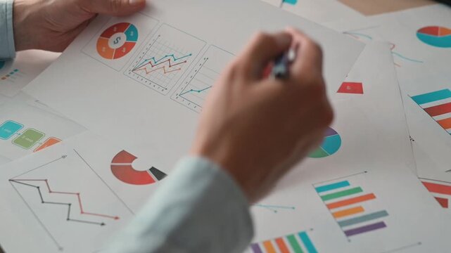 In an office setting, a person is reviewing various charts and graphs on printed documents. The individual uses a pen to mark important data for analysis on multiple sheets of paper