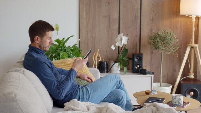 Man sitting on sofa and using smartphone in modern living room. Person holding mobile phone in home interior with plants, lamp and coffee table. Home lifestyle and digital technology concept