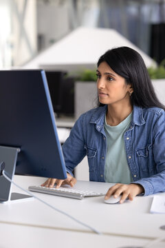 Young Indian woman analyst or marketer sitting at modern workstation, focused on large desktop screen using keyboard and mouse, reviewing data or dashboards, analyzing reports, working on presentation