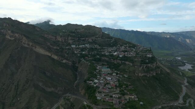 Drone footage of Gunib aul on a high mountain plateau with winding roads, steep cliffs and green valleys, dramatic clouds and summer light, Dagestan, Russia