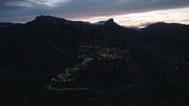 Drone footage at dusk over Gunib village with glowing lights, steep mountain ridges and serpentine roads in Dagestan, Russia, dramatic clouds and sunset horizon