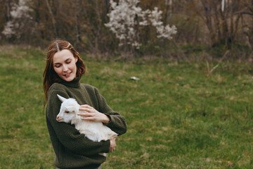Naklejka premium Gentle woman in a dark green sweater holds a white goat kid in a sunlit field, smiling softly, conveying calm rural lifestyle and close animal bond with open meadow behind, gentle breeze