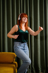 Fototapeta premium Happy red-haired woman dancing with closed eyes in headphones at home, energetic music lifestyle and stress relief concept, joyful girl enjoying favorite song indoors, emotional freedom, domestic fun
