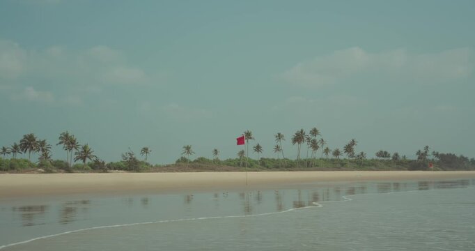 Tropical beach featuring palm trees along the extensive shoreline, a red warning flag flying under a wide blue sky with clouds, reflections of the landscape in the wet sand