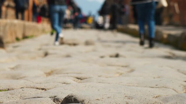 Ground-level shot of unrecognizable tourists walking along Pompeii&rsquo;s ancient stone street, highlighting Roman paving, soft blur, and popular heritage tourism in Italy.