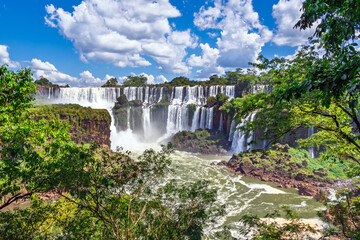 Fototapeta premium Panoramic view of the majestic Iguazu Falls surrounded by lush green tropical rainforest under a blue cloudy sky, Misiones, Argentina