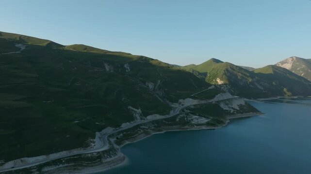 Drone footage of Kezenoyam Lake with winding road along the shore and green mountain ridges in the Caucasus, Chechnya Republic, Russia, on a clear day
