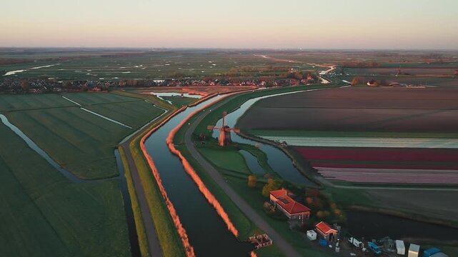 Aerial view of a traditional windmill standing by a canal near colorful tulip fields at sunset in Schermerhorn, North Holland, Netherlands.