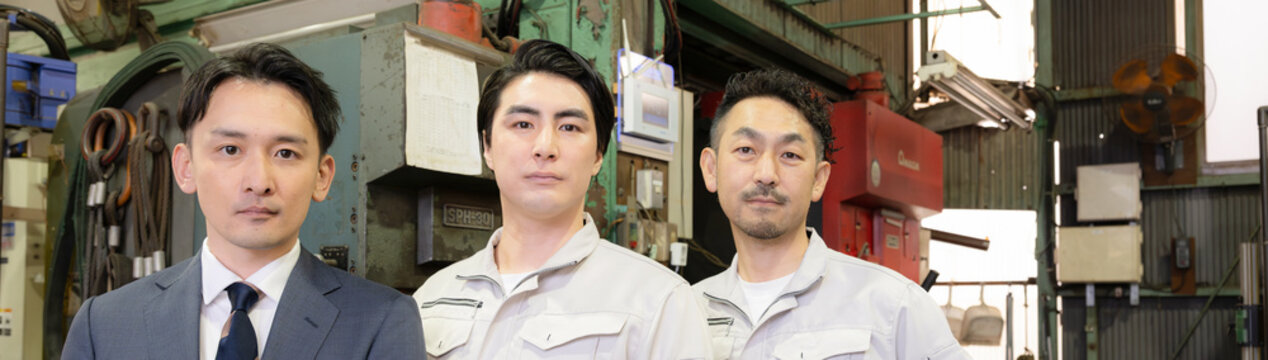 工場の製造業の現場で働く作業着とスーツの営業マン3人チームのポートレート　中小企業のカメラ目線の真剣な表情バナー