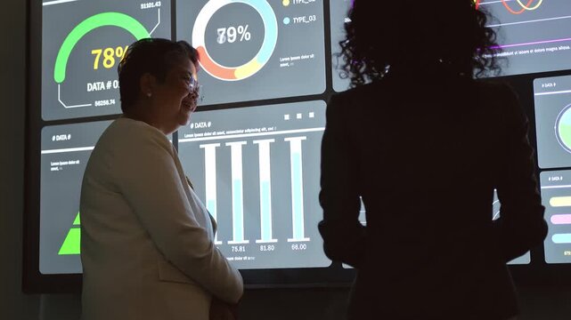 Two professionals review interactive dashboards on a large screen, discussing analytics, charts, and performance metrics in a dim conference room