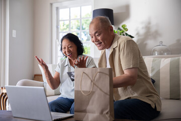 Obraz premium Senior Chinese woman and Chinese man sitting on sofa, gesturing at laptop near paper bag
