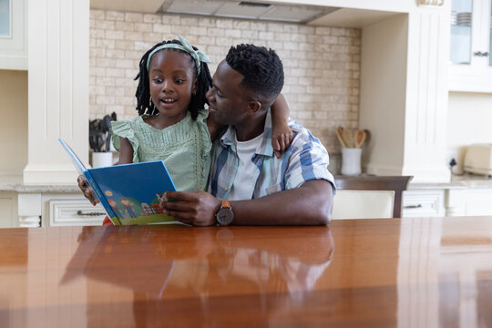 African American father and daughter child reading kids book at home table, plaid shirt green dress