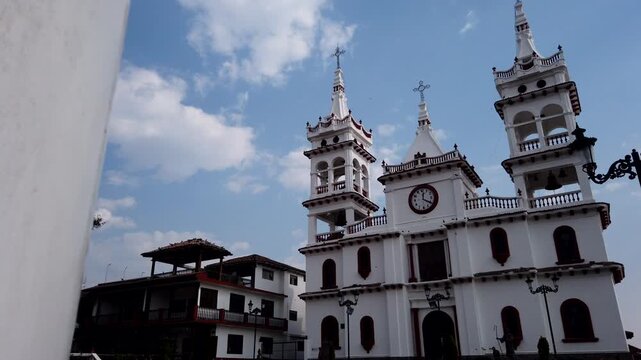 San Crist&oacute;bal parish with white colonial facade, twin bell towers, central clock and nearby rustic buildings near Mazamitla town square, Jalisco, Mexico. Timelapse pan right to left.