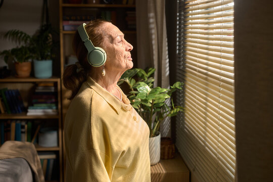 Senior Caucasian woman wearing headphones, standing by window and listening mindfully, representing relaxation and self care. Useful for wellness, retirement, mental health campaigns