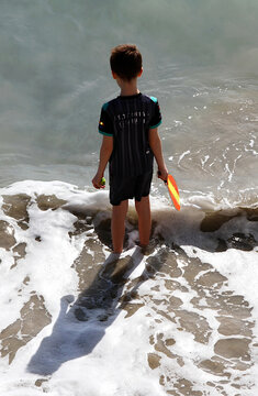 Ni&ntilde;o en orilla de mar para ba&ntilde;o sea con olas de playa, pies en el agua y espuma rompeolas con raqueta y pelota jugando en arena de vacaciones de verano bajo el sol on summer holiday y chapuz&oacute;n