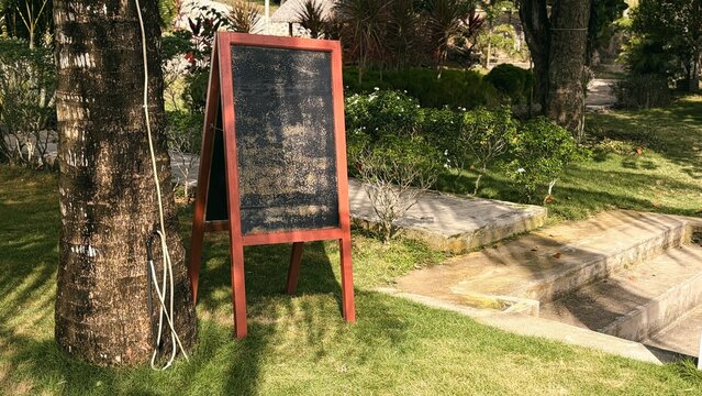 Blank wooden A-frame chalkboard sign on grass in sunny tropical garden, ready for custom message or menu.