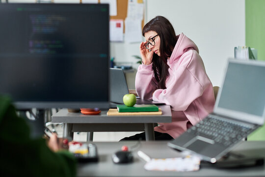 Young adult Caucasian woman working on laptop in office, touching glasses and concentrating on coding task, representing focused tech workflow. For software development or IT career marketing
