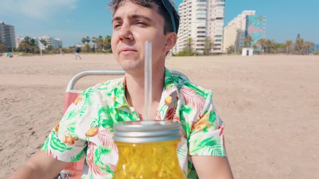 Close up of a young man in a hawaiian shirt drinking a refreshing beverage from a pineapple shaped glass, enjoying his summer vacation while sitting on a chair on a sandy beach