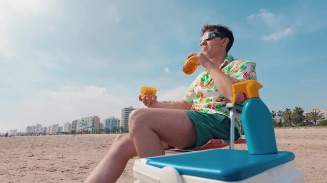 Funny man wearing special protective glasses eating chips and enjoying a drink while watching a solar eclipse on a sandy beach during his summer vacation, with a cityscape in the background