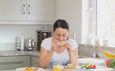 Adult woman sitting at kitchen counter eating sandwich with orange juice and eggs near window light © wavebreak3