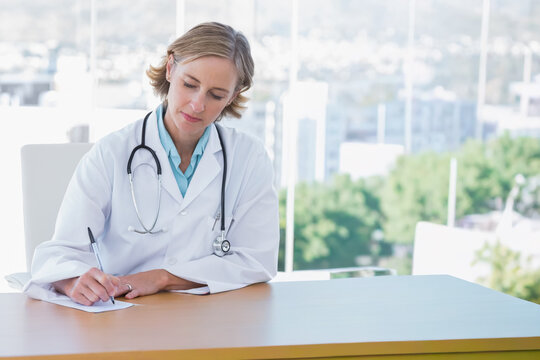 Mature female doctor wearing white lab coat writing on paper at clinic desk with stethoscope
