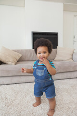Naklejka premium African American infant boy standing, balancing on carpet in living room, wearing denim overalls