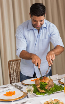 Mid-adult man standing at table carving roasted bird with knife-and-fork wearing light blue shirt