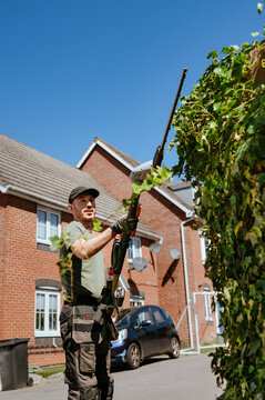 Man trimming tall hedge with electric trimmer in suburban street