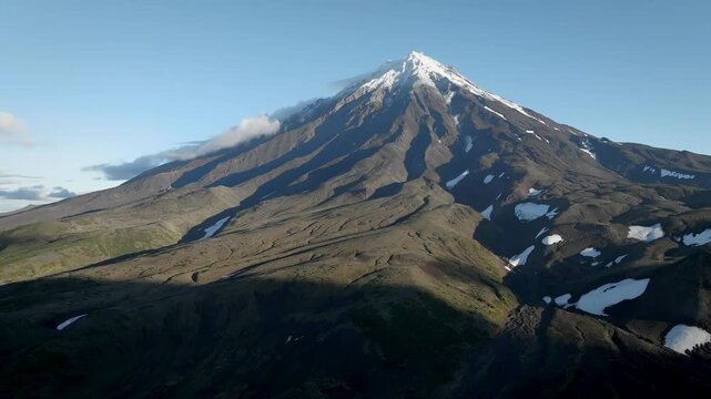Drone footage of Koryaksky Volcano rising above rugged volcanic slopes with lingering snowfields under clear blue sky near Petropavlovsk-Kamchatsky, Kamchatka Krai, Russia