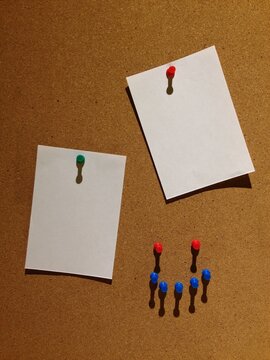 A cork bulletin board with two blank white notes pinned using red and green pushpins, surrounded by extra pins.
