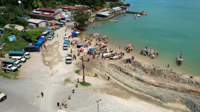 Aerial view of local people gathered near outrigger boats and parked vehicles along the turquoise coastline and tropical greenery in Wewak, East Sepik Province, Papua New Guinea.