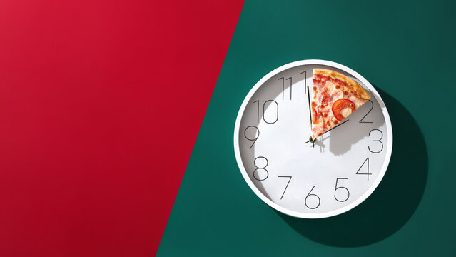 A slice of pizza instead of clock hands on a white wall clock on a red green background. Creative concept pizza time with copy space.