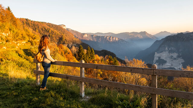 Woman traveler enjoying panoramic autumn mountain view from a viewpoint with a wooden fence, surrounded by vibrant foliage and misty peaks, promoting nature tourism in Luserna, Trentino, Italy.