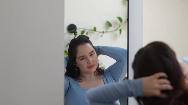Satisfied young woman looking at home mirror with soft smile, feeling confident and self-accepting. Concept of self-love, body positivity and authentic beauty without filters. Inner harmony of female.