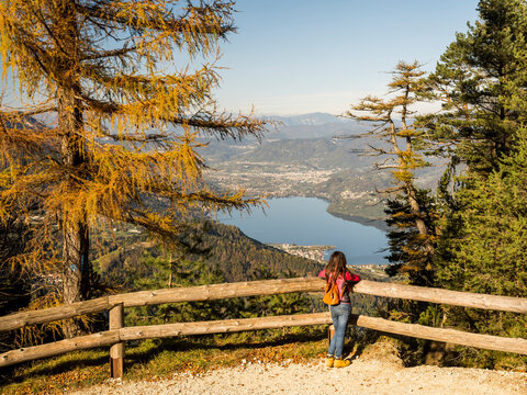 Traveler woman views autumn mountain lake and town panorama from a wooden fence, inspiring outdoor lifestyle and exploration. Respiro degli alberi path, Lavarone, Paganella, Trentino, Italy.