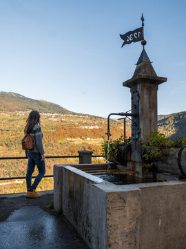 Woman traveler standing by a traditional stone fountain, looking at the scenic autumn mountain landscape, promoting outdoor lifestyle and exploration in Guardia, Murales village, Trentino, Italy.