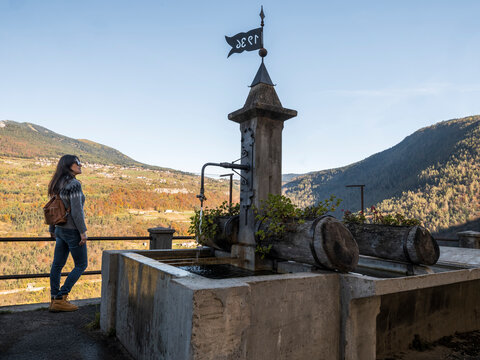 Woman traveler stands by a traditional fountain, admiring the autumn mountain landscape in Guardia, Murales village, Folgaria, Trentino, Italy, a lifestyle travel destination.