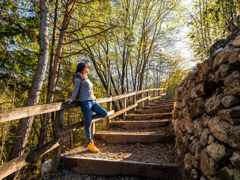 Travel lifestyle woman relaxing on a scenic autumn path, leaning on a wooden railing amidst colorful trees and steps in Guardia, Murales village, Folgaria, Paganella, Trentino, Italy.