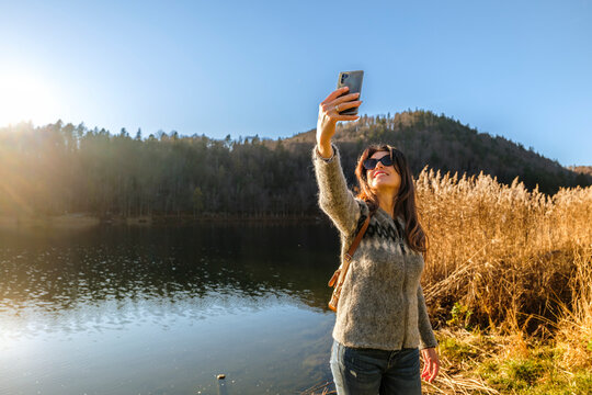 Outdoor travel woman taking a selfie with smartphone by Lavarone lake, surrounded by reeds and forest under a clear sky with sun flare, enjoying relaxation in Trentino, Italy.