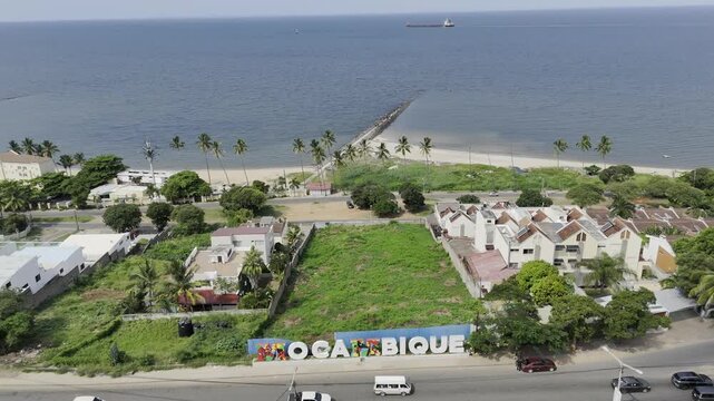 Drone flies over Mozambique photo op sign, toward the sea on sunny day in Maputo, Mozambique